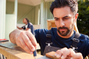 Male Carpenter With Female Apprentice Measuring Wood To Build Outdoor Summerhouse In Garden